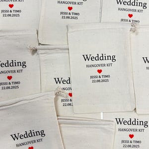May include: Off-white canvas bags with drawstring closures, printed with "Wedding HANGOVER KIT," a red heart, and the names "Jessi & Timo" with the date "22.08.2025." The bags are arranged in a slightly overlapping pattern.