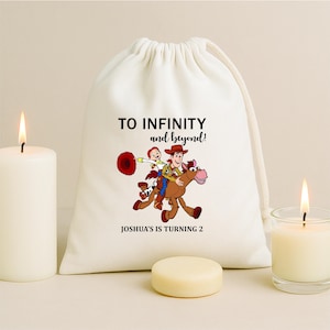 May include: A white drawstring bag with the text "TO INFINITY and beyond!" and "JOSHUA'S IS TURNING 2". The bag features a cartoon illustration of Woody and Buzz Lightyear riding a horse. Two lit candles and a small, round, light-coloured object are in the foreground.