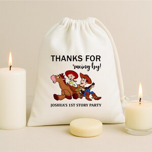 May include: A white drawstring bag with the text "THANKS FOR racing by!" and "JOSHUA'S 1ST STORY PARTY". The bag features a cartoon illustration of two characters riding a horse. A lit candle and a small, round, light-colored object are in the foreground.