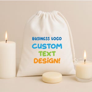 Pode incluir: Uma bolsa branca com cordão com o texto "BUSINESS LOGO CUSTOM TEXT DESIGN!" em azul, verde e laranja. Uma vela acesa e um pequeno objeto redondo e de cor clara estão em frente à bolsa. Uma segunda vela num frasco de vidro está à direita.