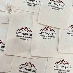 May include: Several off-white canvas pouches are displayed. Each pouch has a dark red mountain range graphic and the text "ALTITUDE KIT" above "DEVINE IN DURANGO 08.09.2025" in black. The pouches are closed with a drawstring.