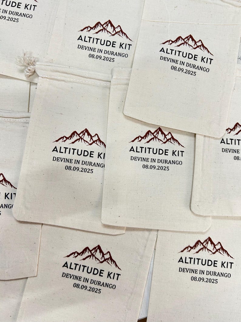 May include: Several off-white canvas pouches are displayed. Each pouch has a dark red mountain range graphic and the text "ALTITUDE KIT" above "DEVINE IN DURANGO 08.09.2025" in black. The pouches are closed with a drawstring.