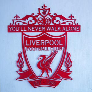 May include: A red metal Liverpool Football Club emblem. The design includes the words "YOU'LL NEVER WALK ALONE" and the club's name. A bird emblem is central, with the text "EST - 1892" at the base. The emblem is a shield shape.