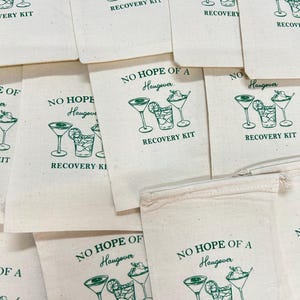 May include: Off-white canvas bags with a green illustration of cocktails and the text "NO HOPE OF A Hangover RECOVERY KIT". Several bags are visible, arranged in a pile.