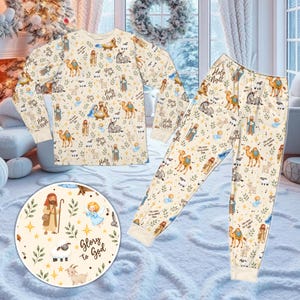 Puede incluir: Un conjunto de pijama de pantalón y camisa de manga larga en color crema con un patrón repetitivo de figuras del nacimiento, camellos, ovejas y ángeles. El estampado incluye el texto Glory to God, Holy Night y Oh come let us adore him.