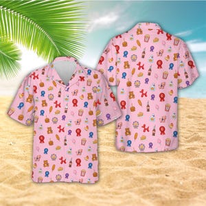 Op de afbeelding: Roze shirt met korte mouwen met een herhaald patroon van illustraties met een carnavals thema. Het patroon bevat teddyberen, linten, hotdogs en andere kermis iconen. Het shirt wordt getoond op een zandstrand.