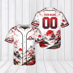 Peut inclure: Un maillot de baseball blanc avec un imprimé paysage rouge et noir représentant des cerisiers en fleurs, des pagodes et un soleil rouge. Le devant affiche le texte ARIRANG, tandis que le dos porte le texte YOUR NAME au-dessus du numéro 00.