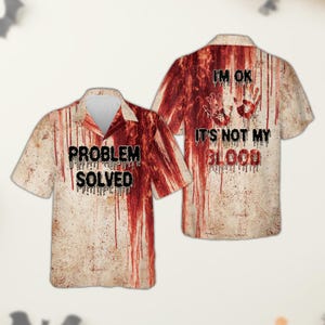 Puede incluir: Una camisa de manga corta con botones, fondo beige y patrones de salpicaduras de sangre roja. El frente muestra el texto "PROBLEM SOLVED" en negro, mientras que la parte posterior presenta el texto "I'M OK IT'S NOT MY BLOOD" con dos huellas de manos rojas.