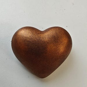 Könnte beinhalten: Ein bronzefarbenes herzförmiges Deko-Objekt. Das Herz hat eine strukturierte Oberfläche und eine abgerundete Form. Das Objekt besteht wahrscheinlich aus Metall oder einem ähnlichen Material. Das Herz ist ein Symbol für Liebe und Zuneigung.