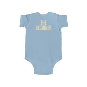 May include: Light blue baby bodysuit with the text "THE BEGINNER" printed in a light beige color across the chest. The bodysuit is a one-piece garment with short sleeves and a round neckline, designed for infants.