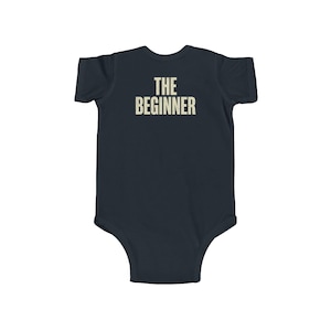 May include: Navy blue baby onesie with the text "THE BEGINNER" printed in a vintage-style font. The onesie is a one-piece garment with short sleeves and a round neckline, designed for infants.