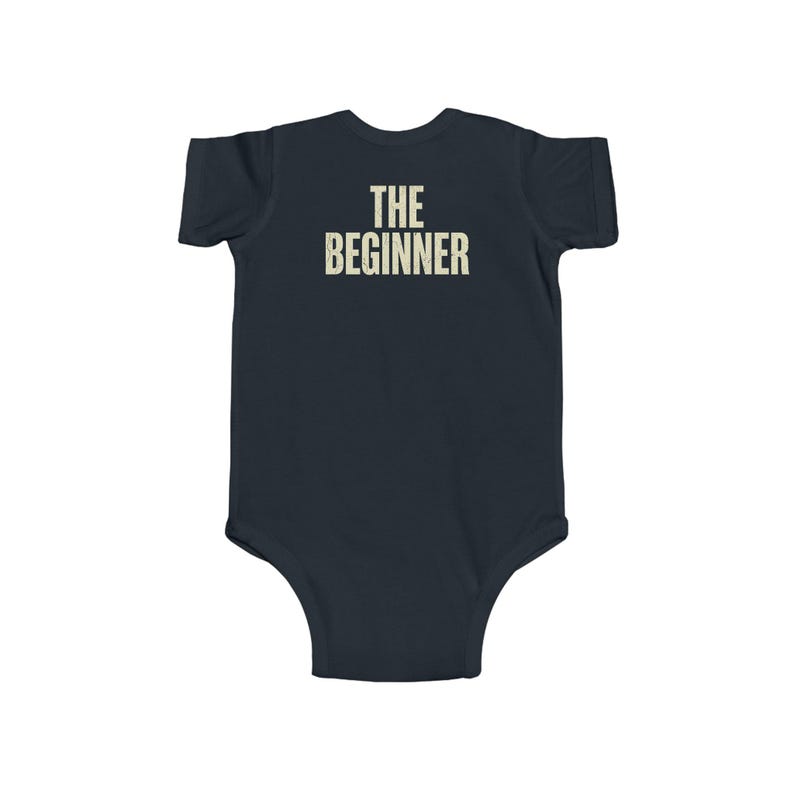 May include: Navy blue baby onesie with the text "THE BEGINNER" printed in a vintage-style font. The onesie is a one-piece garment with short sleeves and a round neckline, designed for infants.
