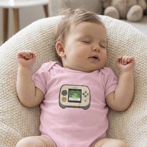 May include: A sleeping baby wearing a light pink onesie with a video game console graphic. The baby is lying on a cream-colored, textured surface. A stuffed bunny and framed photo are in the background.