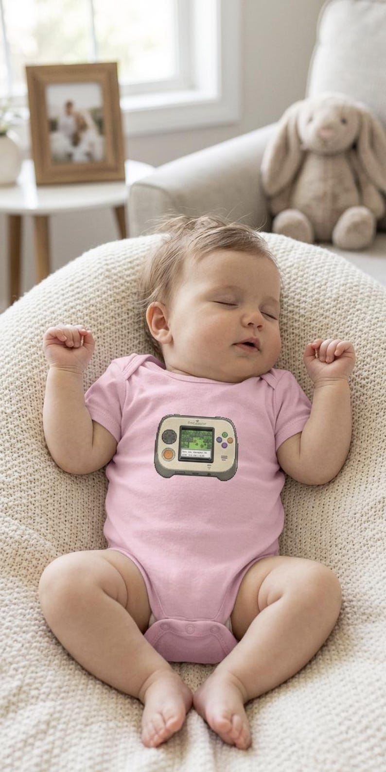 May include: A sleeping baby wearing a light pink onesie with a video game console graphic. The baby is lying on a cream-colored, textured surface. A stuffed bunny and framed photo are in the background.