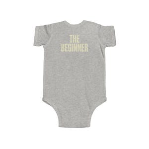May include: A gray baby onesie with short sleeves. The text "THE BEGINNER" is printed in a light tan color across the chest. The onesie is made of a soft material and has a simple, classic design.