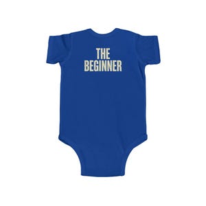 May include: A royal blue baby bodysuit with the text "THE BEGINNER" printed in a light beige color across the back. The bodysuit is short-sleeved and has a round neckline.