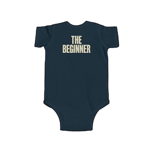 May include: Navy blue baby bodysuit with the text "THE BEGINNER" printed in a vintage, off-white font across the back. The bodysuit is a one-piece garment, designed for infants.