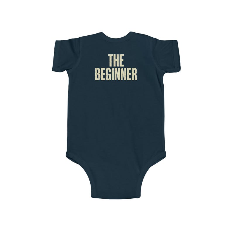 May include: Navy blue baby bodysuit with the text "THE BEGINNER" printed in a vintage, off-white font across the back. The bodysuit is a one-piece garment, designed for infants.