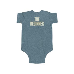 May include: A blue-grey baby onesie with the text "THE BEGINNER" printed in a light beige color across the back. The onesie is made of a soft material and has short sleeves.