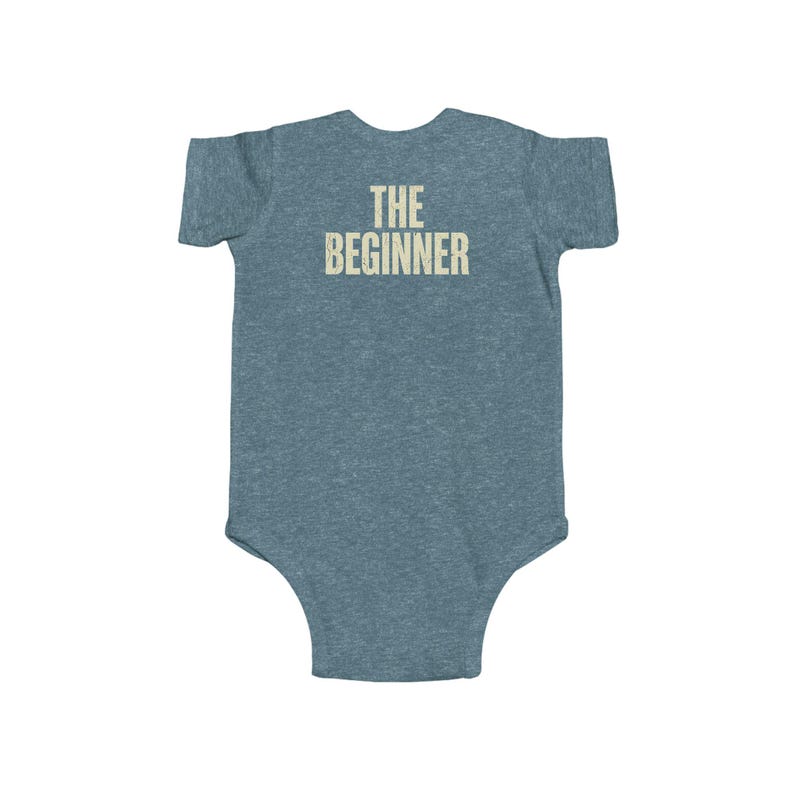 May include: A blue-grey baby onesie with the text "THE BEGINNER" printed in a light beige color across the back. The onesie is made of a soft material and has short sleeves.
