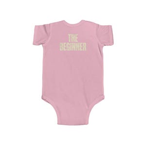 May include: A light pink baby bodysuit with short sleeves. The text "THE BEGINNER" is printed in a light tan color across the chest. The bodysuit is made of a soft material.