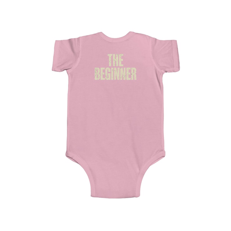 May include: A light pink baby bodysuit with short sleeves. The text "THE BEGINNER" is printed in a light tan color across the chest. The bodysuit is made of a soft material.