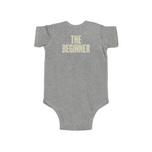 May include: A gray baby onesie with the text "THE BEGINNER" printed in a distressed, light beige font across the back. The onesie is short-sleeved and has a simple, classic design.
