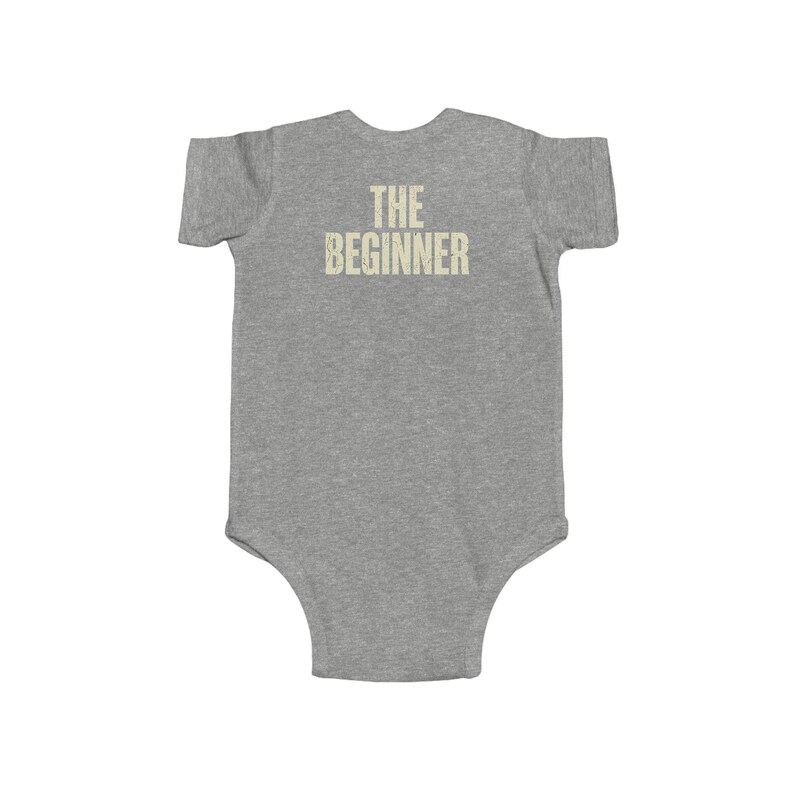 May include: A gray baby onesie with the text "THE BEGINNER" printed in a distressed, light beige font across the back. The onesie is short-sleeved and has a simple, classic design.