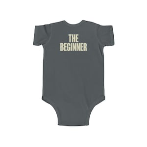 May include: A gray baby bodysuit with the text "THE BEGINNER" printed in a distressed, off-white font across the back. The bodysuit is short-sleeved and has a simple, classic design.