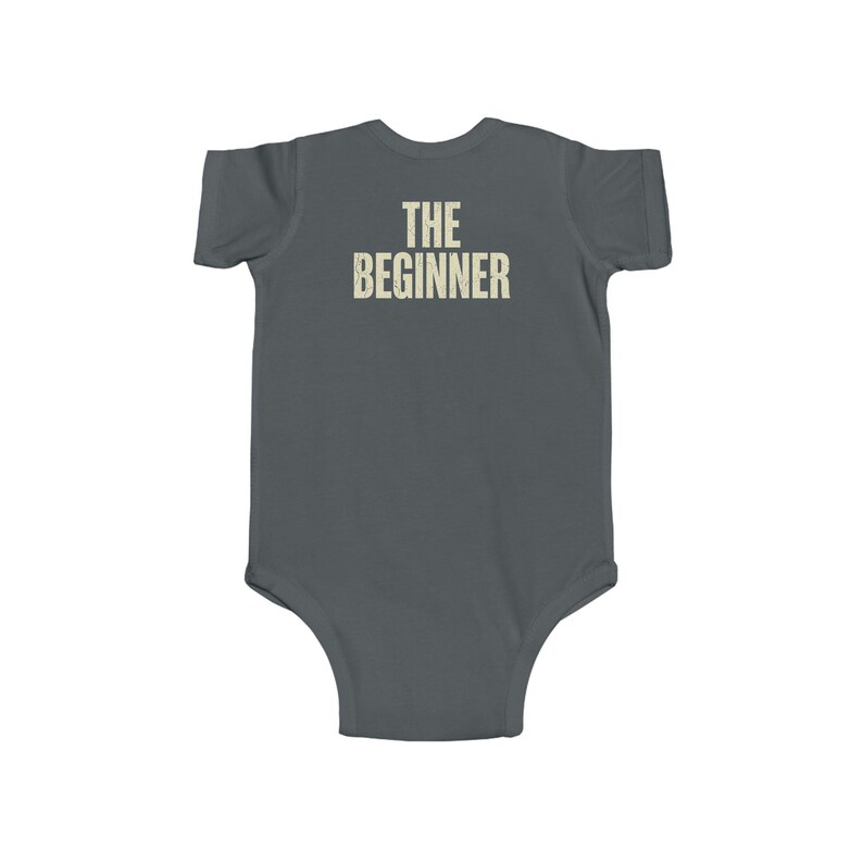May include: A gray baby bodysuit with the text "THE BEGINNER" printed in a distressed, off-white font across the back. The bodysuit is short-sleeved and has a simple, classic design.