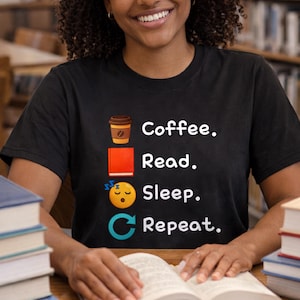 以下が含まれることがあります： 「Coffee. Read. Sleep. Repeat.」の白い文字が入った黒いTシャツ。デザインにはコーヒーカップ、本、眠っている絵文字、円形の矢印が含まれています。本のあるテーブルに人が座っています。