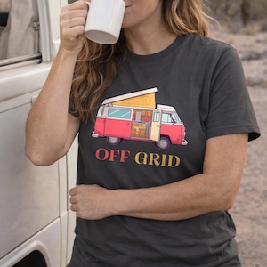 Puede incluir: Camiseta gris oscuro con una ilustración de furgoneta camper roja y blanca y el texto "OFF GRID" en amarillo y rojo. La camiseta es de cuello redondo y la lleva una persona.