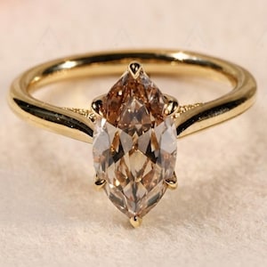 May include: A gold ring featuring a large, marquise-cut champagne-colored gemstone set in a six-prong mounting. The band is smooth and polished, with delicate decorative metalwork visible at the base of the central stone setting.