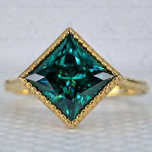 Könnte beinhalten: Ein goldener Ring mit einem quadratisch geschliffenen, smaragdgrünen Edelstein. Der Edelstein ist in einem quadratischen Rahmen mit einem Perlenrand gefasst. Der Ring ist ebenfalls aus Gold, mit den Worten "Emerald Diamonds" im Hintergrund.