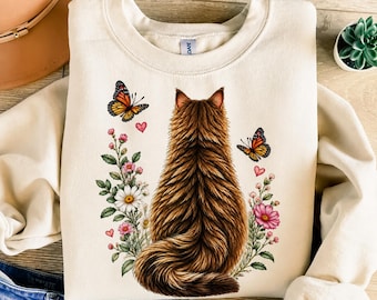Puede incluir: Una sudadera de cuello redondo color crema con un gráfico de un gato marrón esponjoso visto desde atrás. El gato está rodeado de flores silvestres coloridas, hojas verdes, dos mariposas monarca naranjas y pequeños corazones rosas impresos.