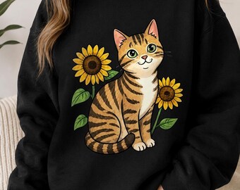 Puede incluir: Una sudadera negra de manga larga con una ilustración gráfica de un gato atigrado marrón con ojos verdes sentado entre dos girasoles amarillos con hojas verdes. El gato tiene rayas de color marrón oscuro y el pecho blanco.