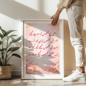 Peut inclure: Impression encadrée avec texte en ourdou rouge, tenue par une personne. L'œuvre présente un fond floral rose et blanc. Le cadre est blanc, et l'impression est rectangulaire.