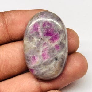 Peut inclure: Une pierre précieuse ovale avec un mélange de couleurs. La pierre présente une base grise et blanche avec des inclusions roses et magenta vibrantes. La surface polie reflète la lumière, soulignant les motifs et les textures naturels de la pierre. Une pièce unique pour la joaillerie.