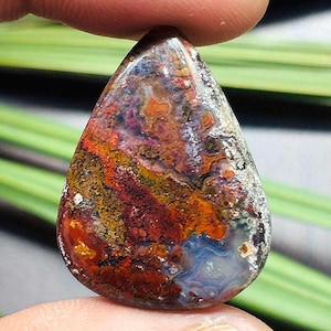 Può includere: Una gemma a forma di goccia con una superficie lucida. La pietra presenta un vivace mix di colori, tra cui rosso, arancione, giallo, blu e bianco. Il motivo assomiglia a un paesaggio con trame vorticose e variazioni di intensità cromatica. La gemma è tenuta tra due dita.