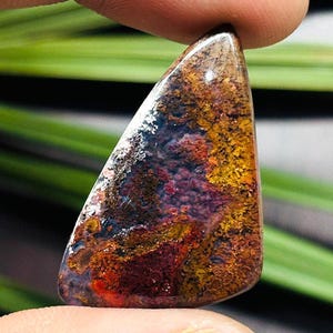 May include: A polished, triangular gemstone with a vibrant mix of colors. The stone features a blend of red, yellow, brown, and white hues, creating a unique, natural pattern. The gemstone is held between two fingers, showcasing its shape and texture.