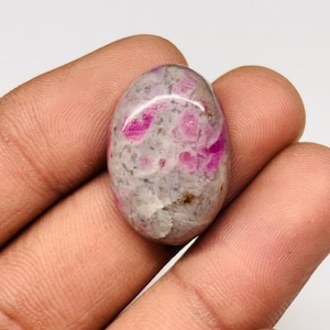 Peut inclure: Pierre précieuse ovale avec un mélange de teintes grises et roses. La pierre présente un motif tacheté de taches roses et magenta, sur une base grise plus claire. La surface polie reflète la lumière, soulignant les textures naturelles de la pierre.