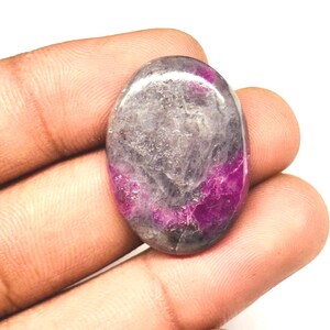 Peut inclure: Une pierre précieuse ovale avec une surface polie. La pierre présente un mélange de couleurs, notamment du gris, du violet et une teinte magenta foncé. La pierre est tenue dans une main, mettant en valeur sa taille et sa forme.