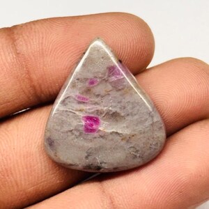 Peut inclure: Une pierre précieuse triangulaire polie, de couleur de base grise avec des inclusions roses vibrantes. La pierre a une surface lisse et brillante, et les taches roses sont dispersées dans la matrice grise. La forme générale est un triangle arrondi.