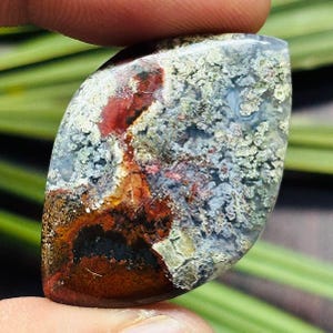 Könnte beinhalten: Ein polierter Edelstein mit einem einzigartigen Farbmuster. Der Stein weist eine Mischung aus roten, braunen, grauen und blauen Farbtönen mit einer strukturierten Oberfläche auf. Die Form ist ein längliches Oval, ideal für Schmuck oder Sammlungen.