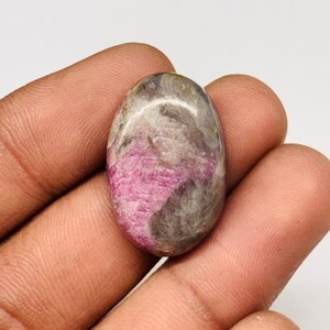 Peut inclure: Une pierre précieuse ovale polie. La pierre présente un mélange de rose, de gris et de brun. Le rose est au centre, tandis que le gris et le brun sont sur les bords. La pierre est tenue dans une main. Idéale pour la joaillerie.