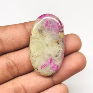 Peut inclure: Une pierre précieuse ovale polie. La pierre présente une base blanc crème avec des stries et des taches roses et rouges. De petits points noirs sont dispersés dans la pierre. La pierre est tenue dans une main.