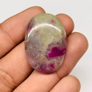 Peut inclure: Une pierre précieuse de forme ovale avec une surface polie. La pierre présente un mélange de couleurs, dont le vert pâle, le gris et le magenta vif. Les teintes magenta sont concentrées en taches irrégulières, créant un motif unique. La pierre est tenue dans une main.