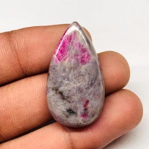 Peut inclure: Une pierre précieuse en forme de goutte polie. La pierre présente un mélange de teintes grises et roses, avec une zone magenta prononcée au sommet. De petites inclusions sombres sont dispersées dans toute la pierre.