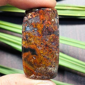 Puede incluir: Una piedra preciosa rectangular con una superficie pulida. La piedra presenta una mezcla de colores, incluyendo rojos profundos, naranjas, amarillos y negros, creando un patrón texturizado único. La piedra preciosa es sostenida por un dedo.