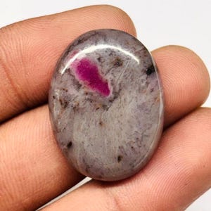 Peut inclure: Pierre précieuse ovale polie. Elle présente un mélange de gris et de blanc, avec une inclusion magenta proéminente. De petits points sombres sont dispersés. Cette pierre unique est idéale pour la création de bijoux.